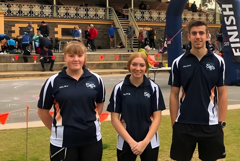 Drei Schüler der Seaton High School tragen Sportsuniformen und stehen vor einem Gebäude mit dekorativer Fassade.