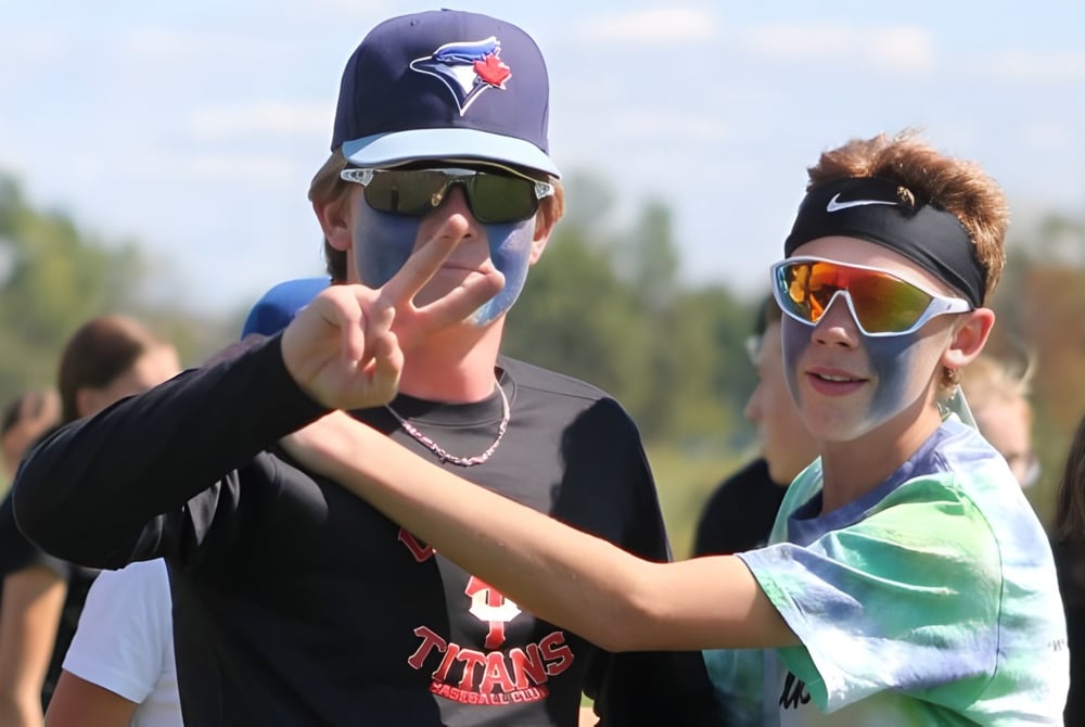 Zwei Personen mit Baseballcaps und Sonnenbrillen machen eine Handgeste vor einem grasbewachsenen Bereich auf dem Gelände der Seaway District High School.