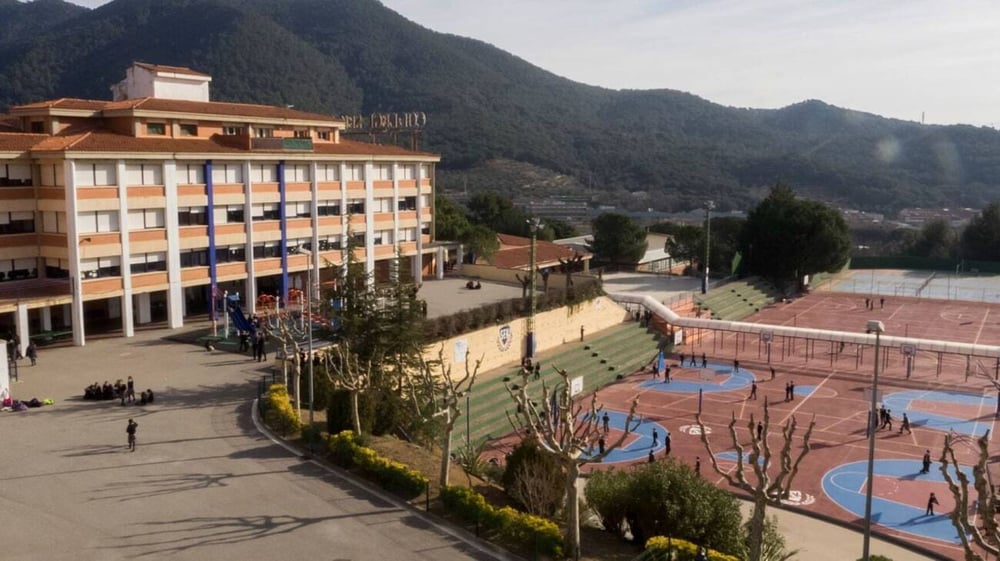 Das mehrstöckige Gebäude der SEK International School Catalunya mit einem bunten Sportplatz im Freien und Bergen im Hintergrund.