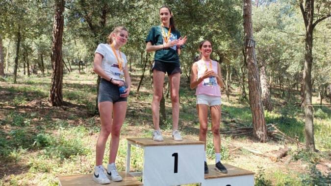 Drei Schülerinnen und Schüler der SEK International School Catalunya stehen auf einem Podium in einem bewaldeten Gebiet, wobei die mittlere Person einen Pokal hält.