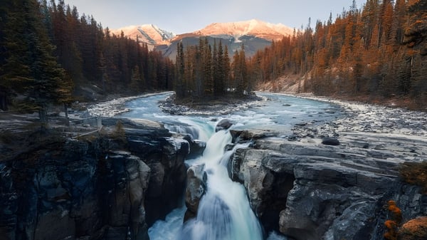 Eine majestätischer Wasserfall fällt über felsige Klippen vor einer Landschaft mit immergrünen Bäumen und schneebedeckten Bergen bei der Semiahmoo Secondary School.