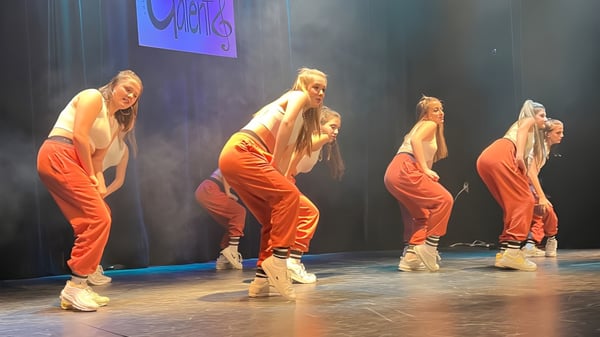 Vier Tänzerinnen und Tänzer in orangen Outfits führen auf der Bühne im Séminaire de Chicoutimi eine Tanzvorführung vor blau-lila Hintergrund auf.