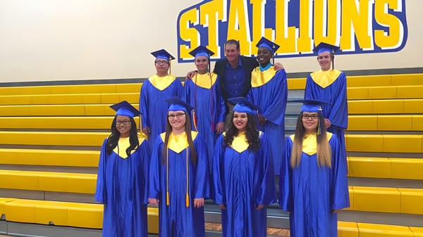 Eine Gruppe Absolventinnen und Absolventen in blauen Talaren steht vor dem Stallions-Schild auf den Gelben Tribünen der Sequoia Charter High School.