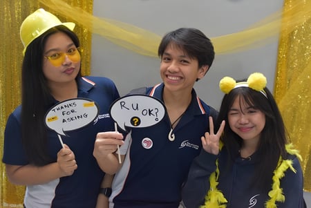 Drei Schülerinnen und Schüler des Sevenoaks Senior College halten Schilder in einem bunten, festlichen Rahmen.