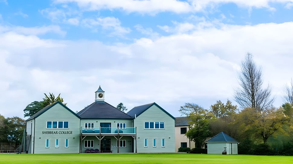 Das zweistöckige Gebäude mit Uhrturm des Shebbear College steht auf einer Wiese mit Bäumen im Hintergrund unter blauem Himmel.