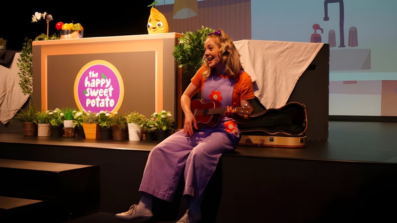 Eine Frau spielt Gitarre auf der Bühne am Sheldon College vor einer Happy Couch Potato Dekoration.