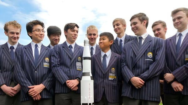 Eine Gruppe junger Männer des Sheldon College steht in dunkelblauen Anzügen zusammen vor einem bewölkten Himmel.