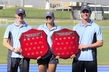 Drei Personen in einheitlicher Kleidung mit roten Schilden stehen auf dem Sportfeld des Sheldon College.