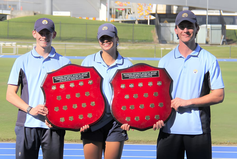 Drei Personen in einheitlicher Kleidung mit roten Schilden stehen auf dem Sportfeld des Sheldon College.