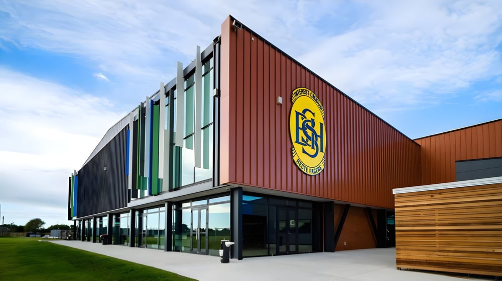 Das mehrfarbige Gebäude mit großem gelbem Logo von Shirley Boys' High School steht auf einem grasbewachsenen Gelände unter blauem Himmel.