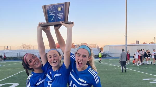 Drei Schülerinnen der Shore Regional High School feiern mit einem Pokal auf dem Sportplatz vor einer Stadtkulisse.