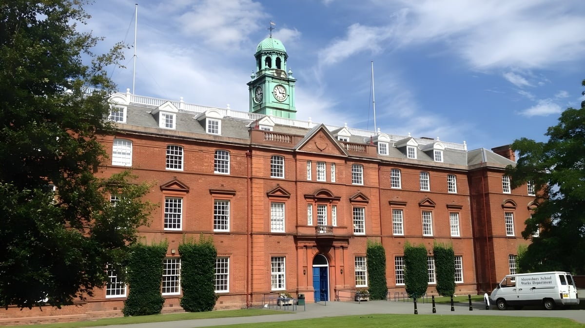 Das historische Backsteingebäude mit grünem Turmdach steht auf dem Campus der Shrewsbury School vor blauem Himmel.