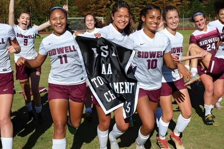Eine Gruppe junger Fußballspielerinnen der Sidwell Friends School versammelt sich lächelnd auf einem grasbewachsenen Spielfeld mit Bäumen im Hintergrund.