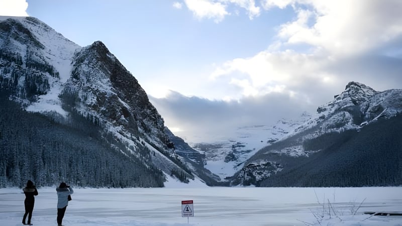 Zwei Personen gehen an einem zugefrorenen See mit schneebedeckten Bergen im Hintergrund nahe der Sir Guy Carleton Secondary School spazieren.