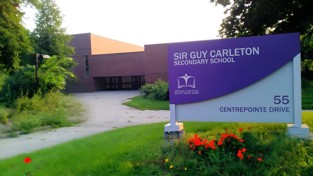 Der Eingang der Sir Guy Carleton Secondary School mit einem großen Schild und einer gepflegten Blumenwiese im Vordergrund.