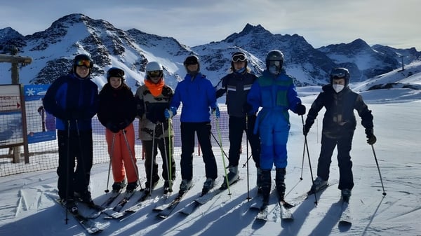 Eine Gruppe von Personen in Winterkleidung steht auf einem schneebedeckten Hang mit schneebedeckten Bergen im Hintergrund auf dem Gelände der Sligo Grammar School.