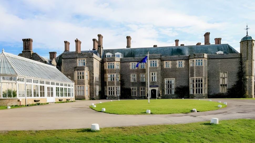 Das historische Herrenhaus mit Gewächshaus und gepflegtem Rasen auf dem Campus des Slindon College bei bewölktem Himmel.