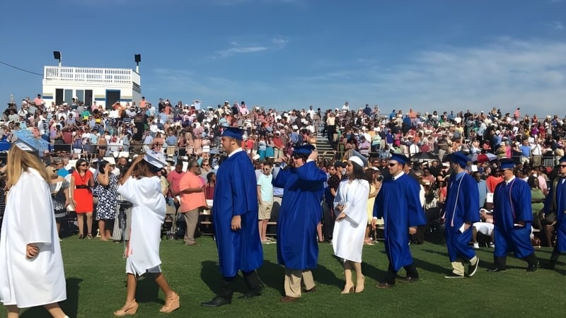 Eine große Gruppe von Absolventinnen und Absolventen der Smithfield State High School steht auf einer Wiese in ihren Talaren mit Hüten unter blauem Himmel.