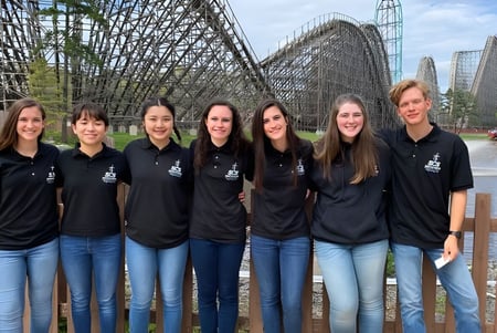 Eine Gruppe von sieben Schülerinnen der Smithtown Christian School steht vor einer großen Achterbahn in einem Freizeitpark.