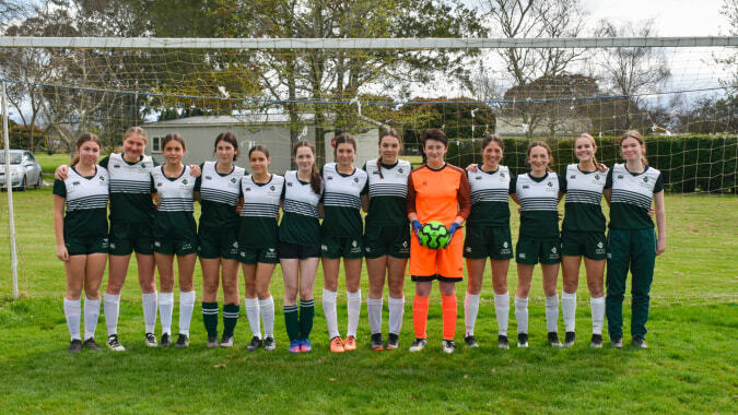 Eine Gruppe junger Fußballspielerinnen steht auf dem Sportplatz des Solway College vor einem Gebäude und Bäumen.