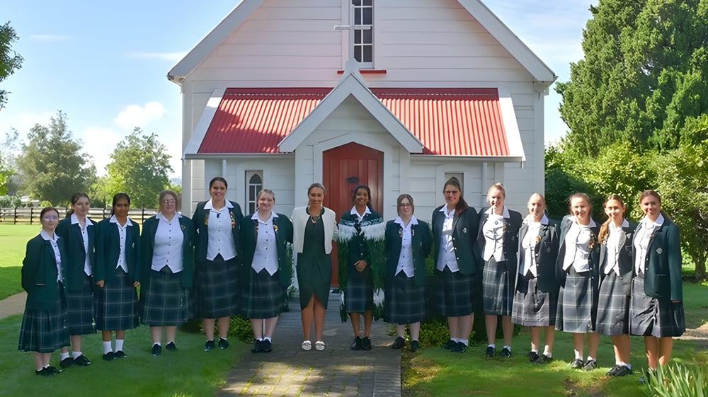 Eine Gruppe junger Schülerinnen in Schuluniformen steht vor einem weißen kirchenähnlichen Gebäude mit rotem Dach auf dem Campus des Solway College.