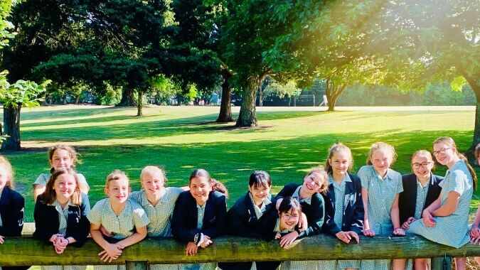 Eine Gruppe junger Schülerinnen des Solway College sitzt auf einer Holzbank im grünen Park mit Bäumen und Sonnenlicht.