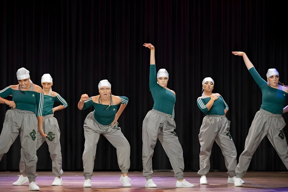Eine Gruppe Schülerinnen und Schüler des Solway College führt auf einer Bühne vor schwarzem Vorhang einen Tanz in grünen und grauen Uniformen auf.