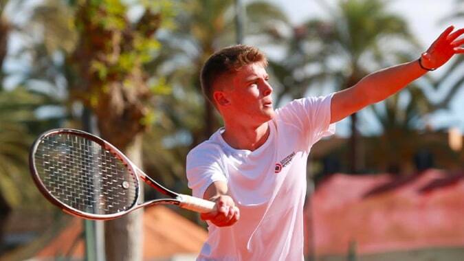 Ein männlicher Tennisspieler in weißem Shirt spielt vor einer Kulisse mit Palmen auf dem Campus der Sotogrande International School.