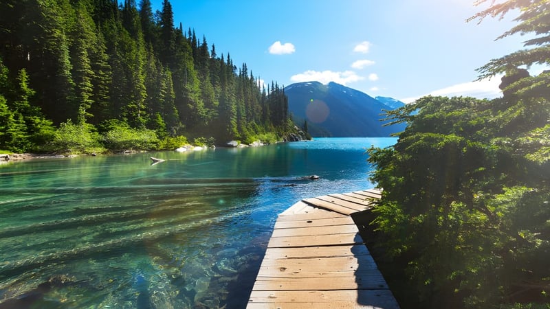 Ein hölzerner Steg erstreckt sich auf einem klaren See in der Nähe der South Delta Secondary School mit Bergen und Wäldern im Hintergrund.