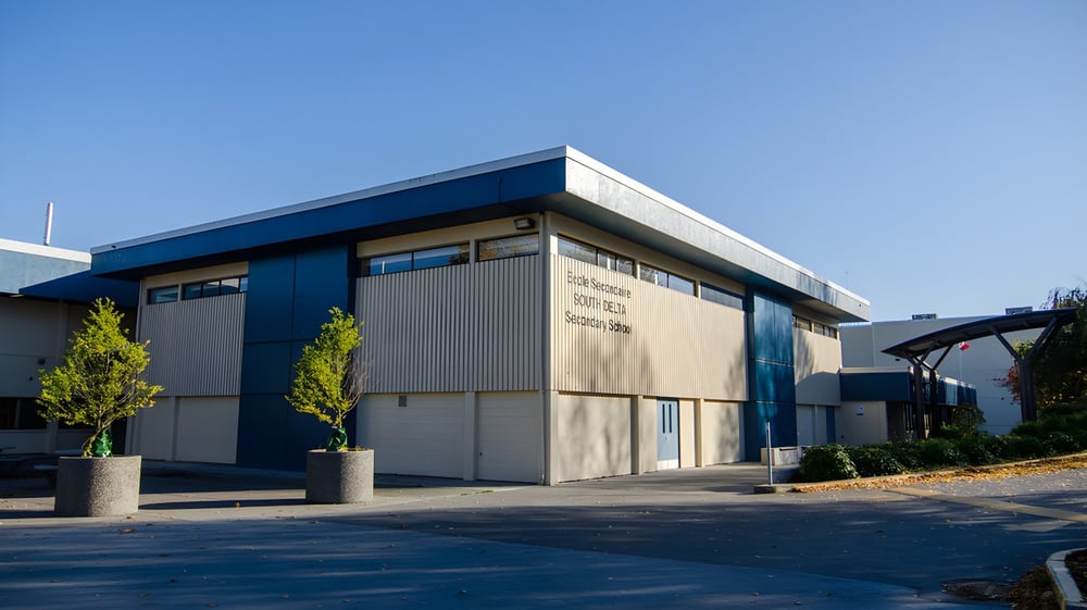 Das moderne Industriegebäude mit blauem Dach auf dem Gelände der South Delta Secondary School steht vor Bäumen und einem gepflasterten Parkplatz.