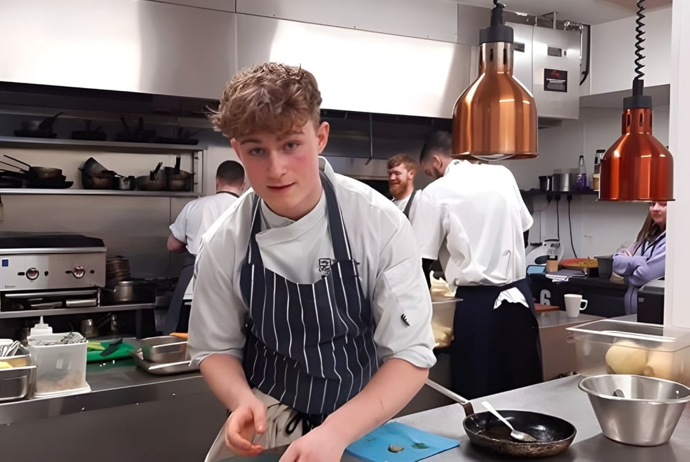Ein Schüler in Kochuniform steht in der Küche auf dem Campus des South Downs College umgeben von Kochgeräten.