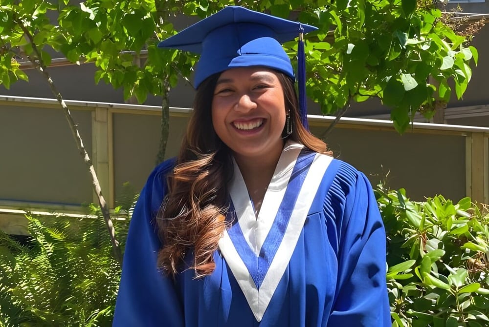 Eine Absolventin im blauen Abschlussgewand steht vor einem grünen Garten auf dem Campus des South Hill Education Centre.