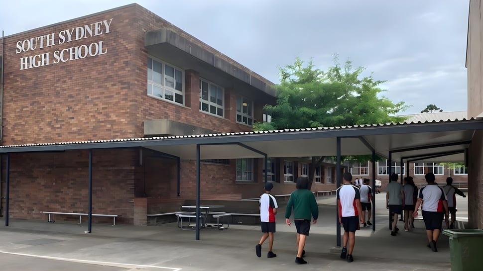 Das Backsteingebäude der South Sydney High School ist von Bäumen umgeben, während Schüler auf dem Gehweg vorbeigehen.