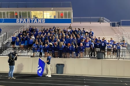 Die Sportmannschaft der Sparta High School versammelt sich auf den Tribünen eines Stadions mit dem Schriftzug Spartans im Hintergrund.