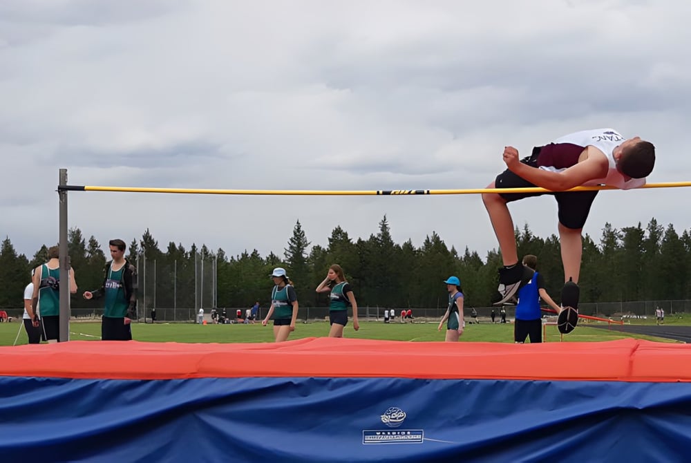 Ein Schüler der Sparwood Secondary School springt im roten Trikot über eine hohe Latte beim Hochsprung im Freien.