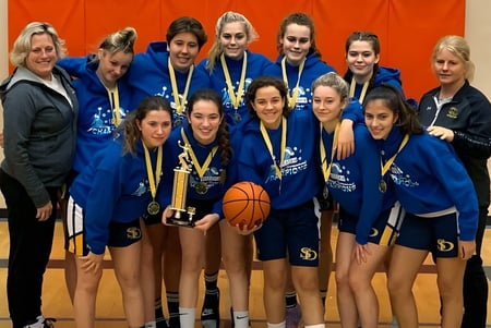 Eine Gruppe weiblicher Basketballspielerinnen der Spectrum Community Secondary School hält in der Sporthalle einen Pokal und einen Basketball.