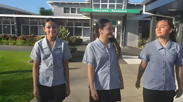 Drei Schülerinnen in grauen Uniformen stehen vor einem Gebäude mit grünem Schild auf dem Campus des Spotswood College.