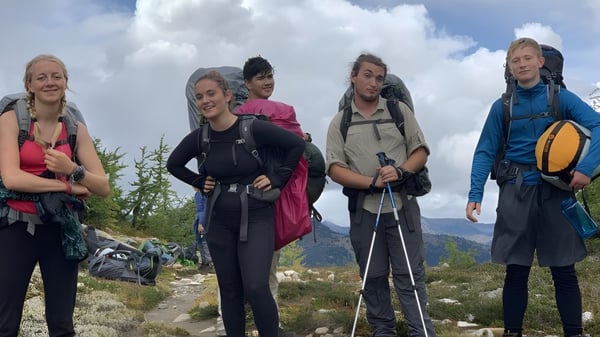 Eine Gruppe Schülerinnen der Spring Street International School steht in Wanderausrüstung in einer bergigen Landschaft mit Wolken und blauem Himmel.