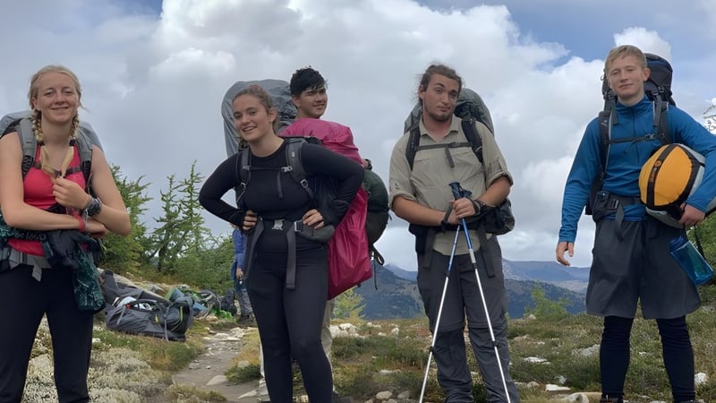 Eine Gruppe Schülerinnen der Spring Street International School steht in Wanderausrüstung in einer bergigen Landschaft mit Wolken und blauem Himmel.