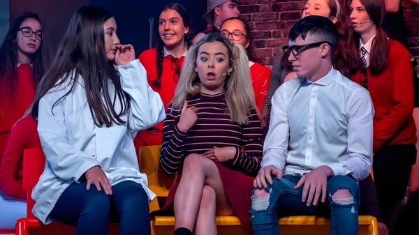 Eine Gruppe Schüler der St. Aidan's Comprehensive School sitzt auf der Bühne vor einer Backsteinwand in einer Aufführungssituation.