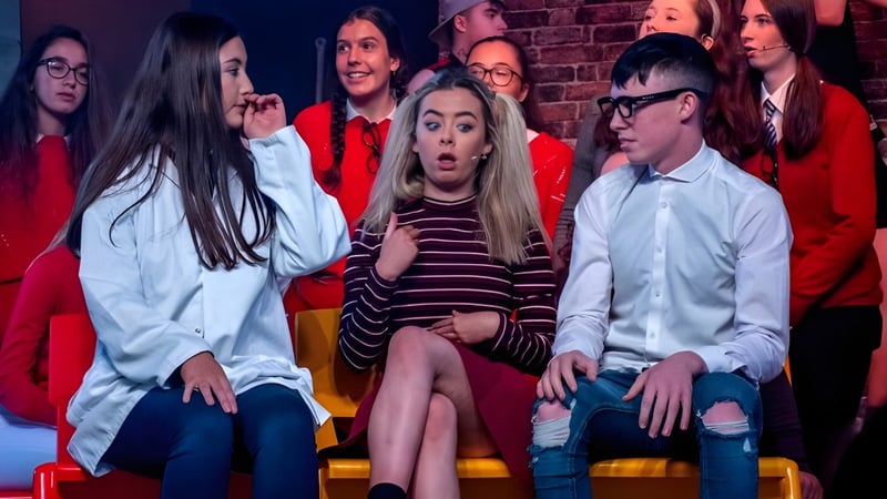 Eine Gruppe Schüler der St. Aidan's Comprehensive School sitzt auf der Bühne vor einer Backsteinwand in einer Aufführungssituation.