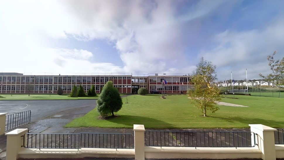 Das Schulgebäude von St. Aidan's Comprehensive School ist hinter einem Zaun mit einer Wiese und Bäumen im Vordergrund unter bewölktem Himmel zu sehen.
