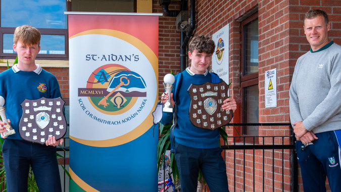 Drei Jungen stehen vor einem Backsteingebäude mit einem farbenfrohen Logo an der Wand der St. Aidan's Comprehensive School.
