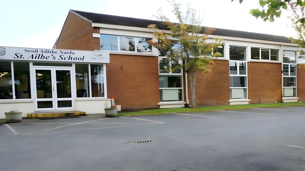 Das zweistöckige Backsteingebäude der St Ailbe's School mit großen Fenstern steht auf einem gepflasterten Gelände umgeben von Bäumen.