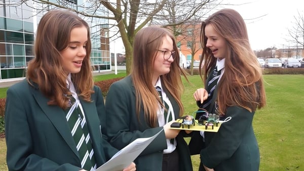 Drei Schülerinnen in Schuluniformen stehen auf dem Gelände der St. Alban's Catholic High School mit Gebäude und Bäumen im Hintergrund.