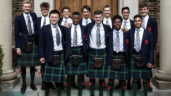 Eine Gruppe Schüler in formeller Kleidung steht vor einer Holztür auf dem Gelände des St. Andrew’s College.