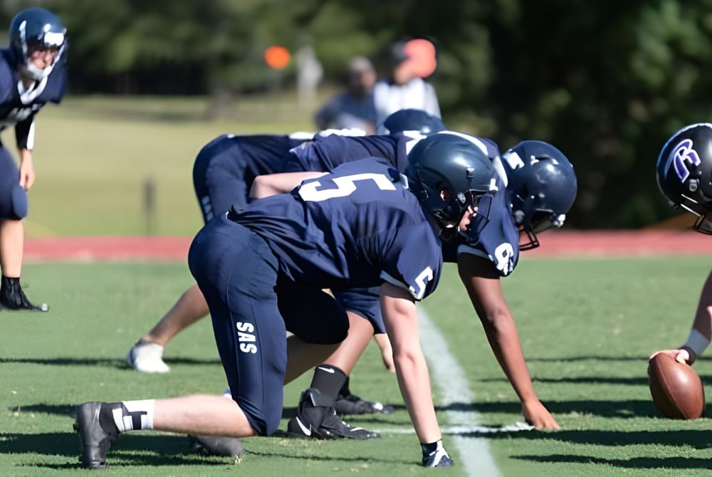 Eine Gruppe von Football-Spielern der St. Andrew's-Sewanee School steht in Verteidigungsposition auf einem Grasfeld.