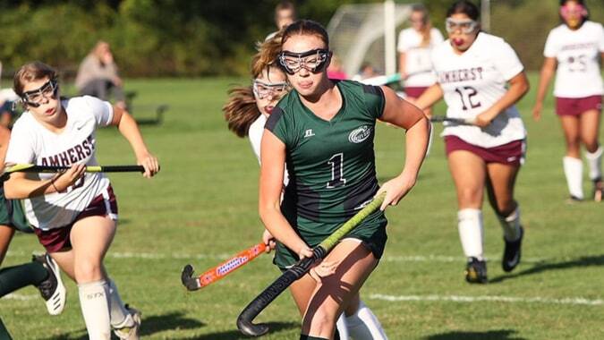 Schülerinnen des St. Angela’s College spielen Feldhockey auf einem Rasenfeld mit Zuschauern im Hintergrund.