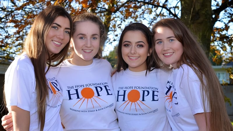 Vier Schülerinnen der St. Anne's Secondary School stehen vor einem Baum mit herbstlichem Laub und tragen weiße T-Shirts mit dem HOPE-Logo.