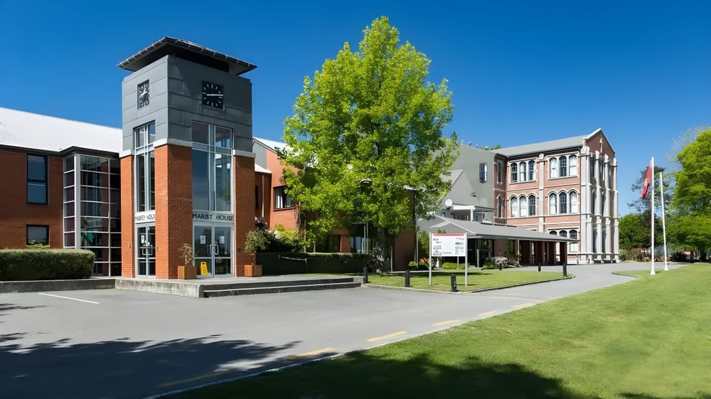 Der hohe Uhrturm auf dem Campus von St. Bede's College ist umgeben von grünen Bäumen und modernen Gebäuden.